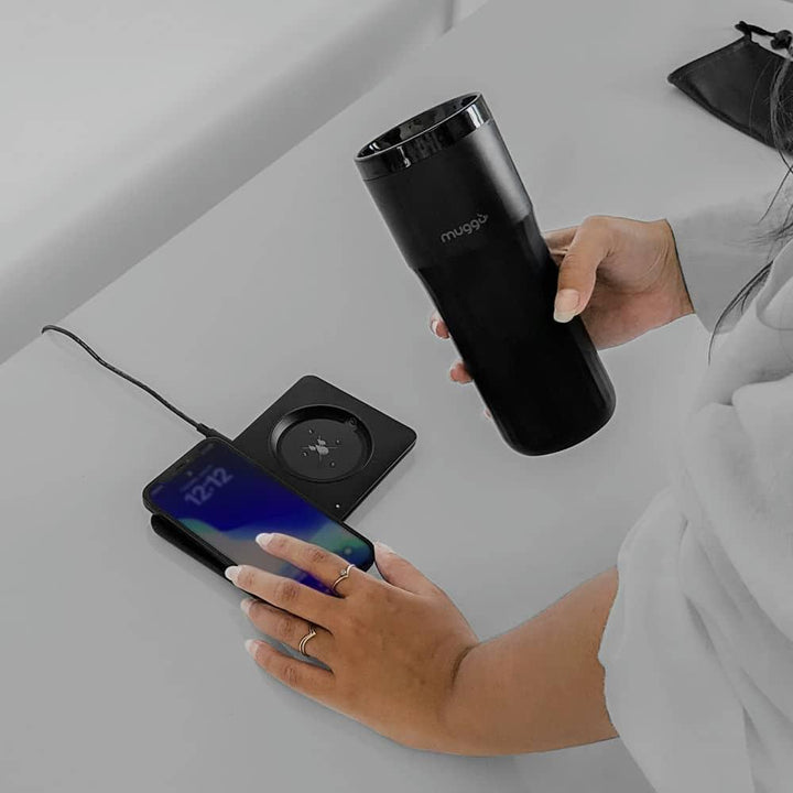 Frau hält schwarzen Muggo Becher neben kabellosem Ladegerät und Smartphone auf Tisch