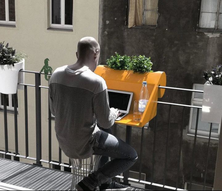 Ein Mann arbeitet an einem Laptop an einem leuchtend gelben Balkontisch mit Topfpflanzen und Blick auf ein Wohngebäude. In der Nähe steht eine Wasserflasche.