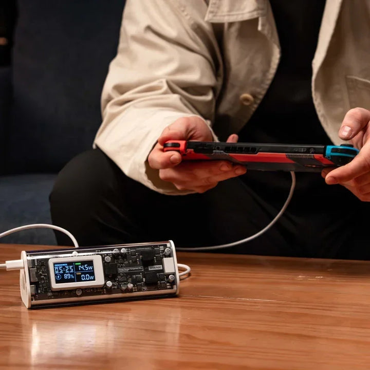 Eine Person lädt eine Spielkonsole mithilfe eines digitalen Lademonitors auf einem Holztisch auf und zeigt die Ladestatistiken des Geräts an.