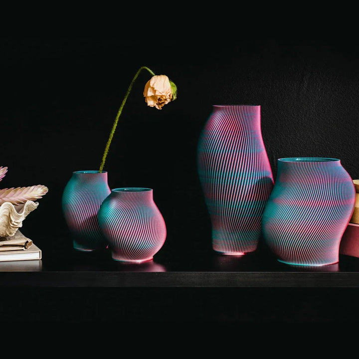 Eine Sammlung lebendiger Vasen mit wellenförmigem Muster in Rosa und Blau mit einem einzelnen Stiel einer getrockneten Blume vor einem schwarzen Hintergrund.