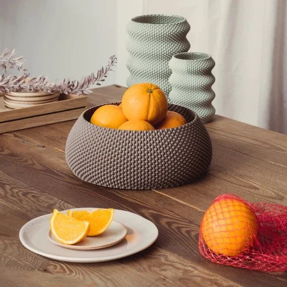 Auf einem Holztisch steht eine strukturierte graue Schüssel voller Orangen, daneben ein Teller mit Orangenscheiben und eine Orange in einem Netzbeutel.