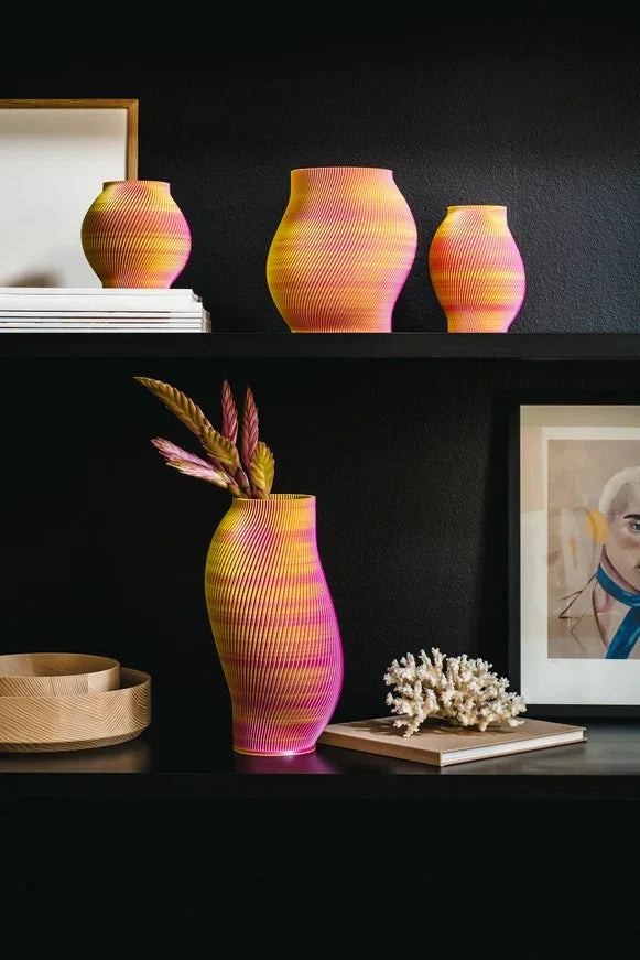 Bunt gestreifte Vasen in Rosa und Gelb mit Blumenarrangement, präsentiert auf einem dunklen Regal neben Kunst- und Dekorationsobjekten.