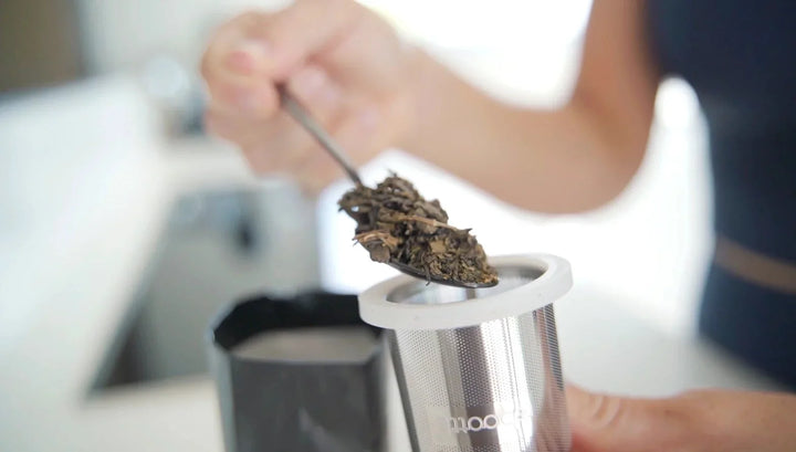 Frau füllt losen Tee mit Löffel in Edelstahl-Teefilter im modernen Küchenumfeld.