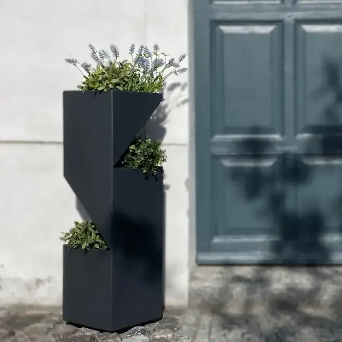 Ein moderner, schwarzer geometrischer Blumenständer mit üppigen grünen Pflanzen und lavendelfarbenen Spitzen, vor einer strukturierten Wand und einer blauen Tür.