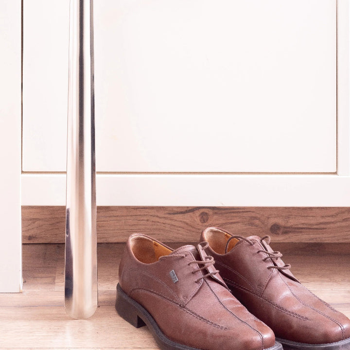 Zwei elegante braune Lederschuhe stehen auf einem Holzboden neben einem glänzenden, metallischen vertikalen Schuhlöffel. Einfache, moderne Inneneinrichtung.