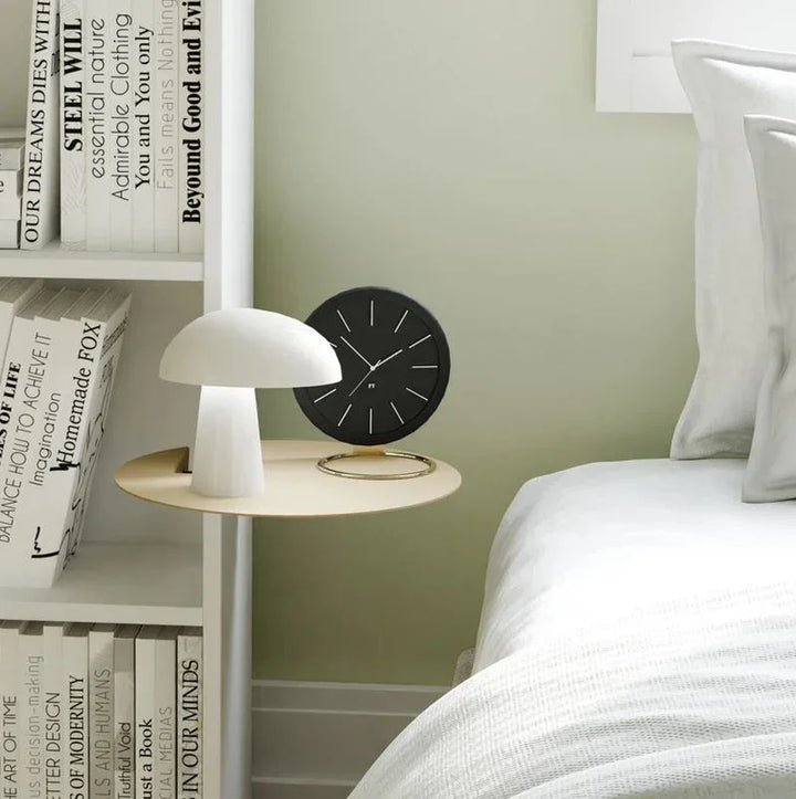 Modernes Schlafzimmer mit weißer Pilzlamp, schwarzer Wanduhr und Bücherregal am Bett.