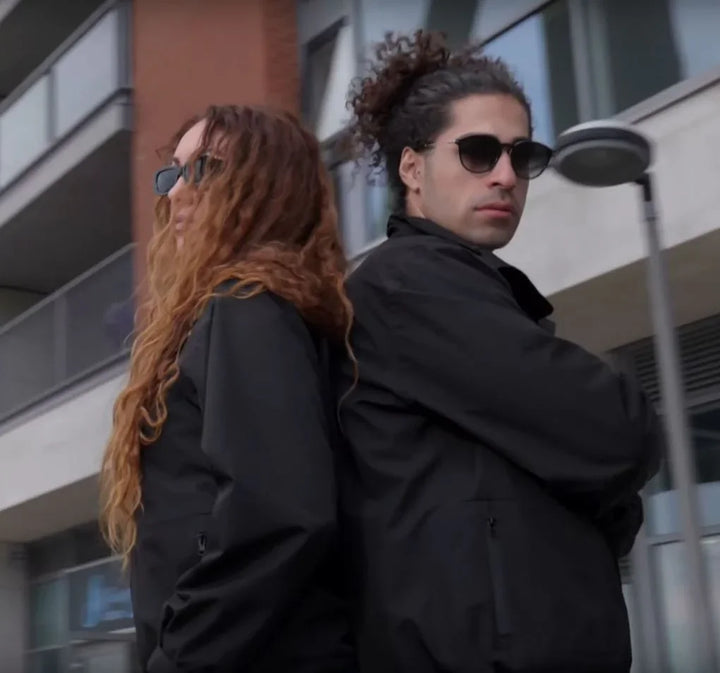 Zwei Personen in schwarzen UZE Jacken und Sonnenbrillen vor modernem Gebäude